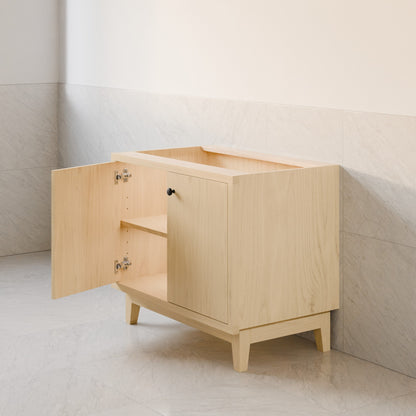 A light natural wood vanity cabinet with one door open, showing a shelf and interior storage space, set against a tiled wall and floor.