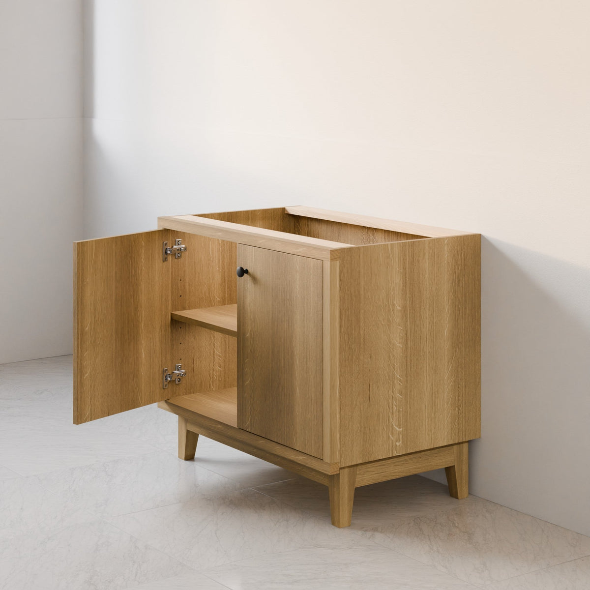A light oak wood storage cabinet with one door open, showing a shelf inside, set against a minimalist off-white wall and light stone floor.