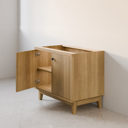 A light oak wood storage cabinet with one door open, showing a shelf inside, set against a minimalist off-white wall and light stone floor.