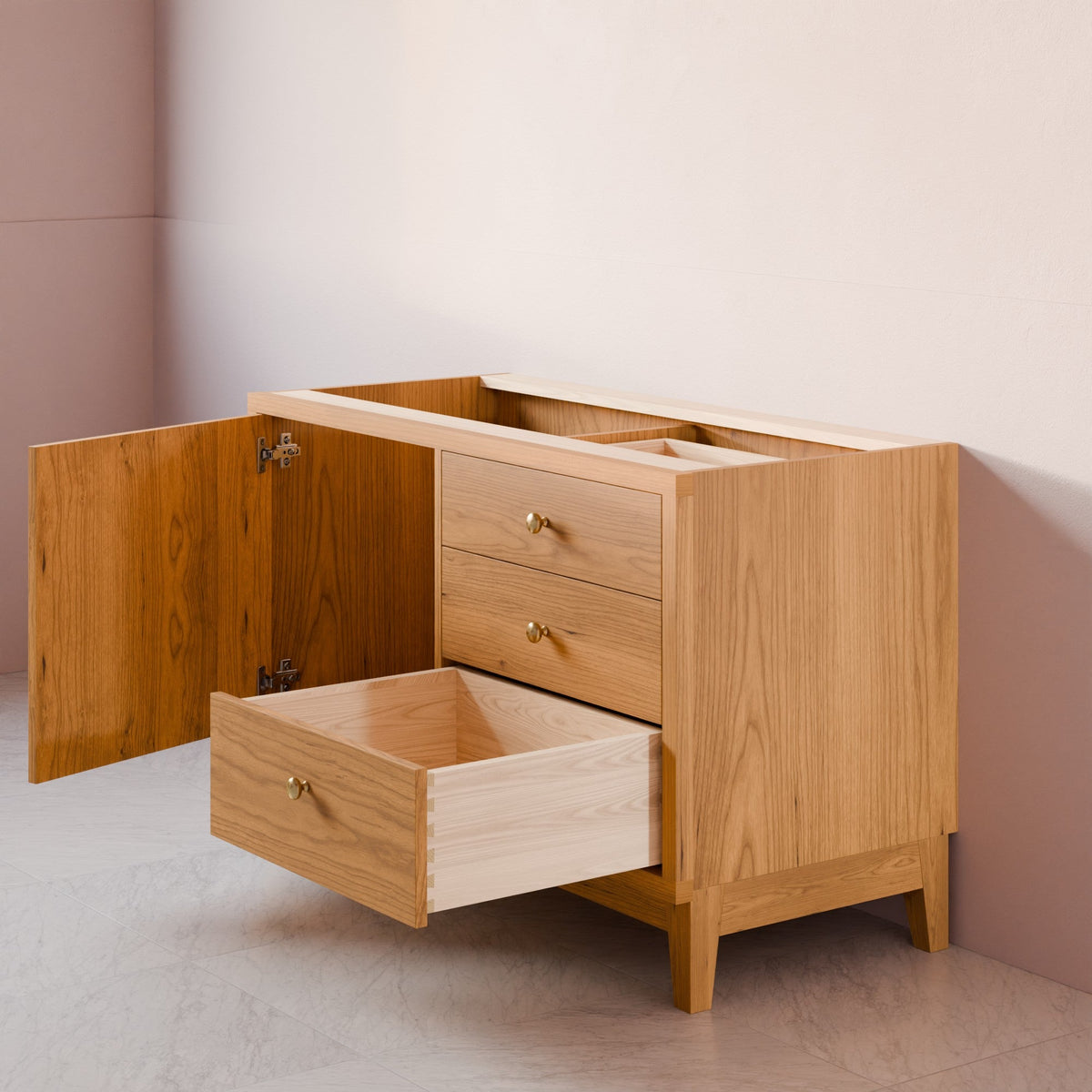 A light oak wood vanity cabinet with an open door and one pulled-out drawer, showing brass hardware, set against a minimalist beige and pink wall background.