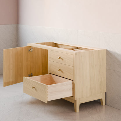 A light natural wood vanity cabinet with three drawers, one open and one pulled out, set against a soft pink and beige tiled wall.