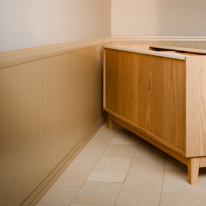 A mid-century modern wooden cabinet with a warm honey finish, set in a corner of a room with beige wainscoting and light stone tile flooring.