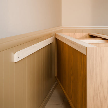 A close-up of a wooden wall-mounted shelf or ledge installed in a beige paneled wall corner next to a wood vanity or cabinet.