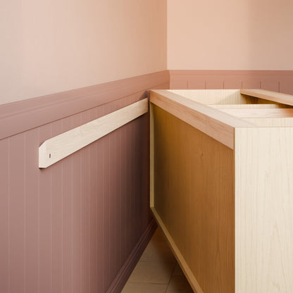 A close-up of a light natural wood wall-mounted shelf or cabinet rail installed on a dusty rose paneled wall next to a light wood cabinet base.