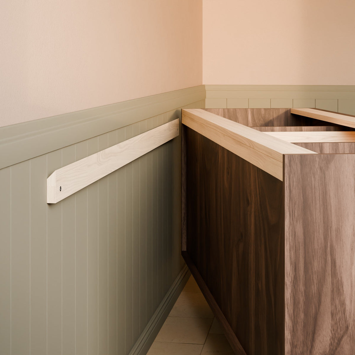 A close-up of a wooden kitchen cabinet corner with a dark wood front, light wood top, and a matching light wood wall-mounted trim piece against a beige and sage green wainscoted wall.