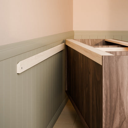 A close-up of a wooden kitchen cabinet corner with a dark wood front, light wood top, and a matching light wood wall-mounted trim piece against a beige and sage green wainscoted wall.
