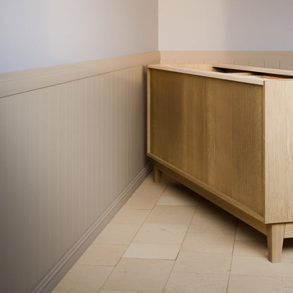 A light oak wood sideboard cabinet in a corner room with beige paneled walls and light stone tile flooring.