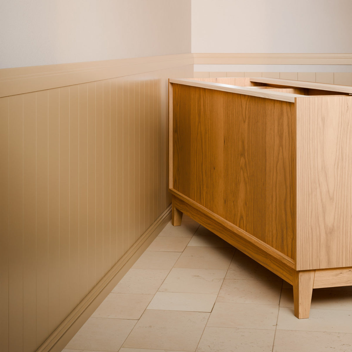 A light natural wood vanity cabinet with a white countertop, shown in a bathroom corner with beige wainscoting and tiled floor.