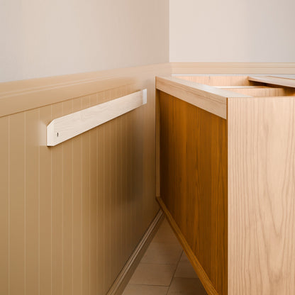 A light natural wood wall-mounted shelf or ledge installed on a beige paneled wall next to a wooden cabinet, shown in a clean interior setting.