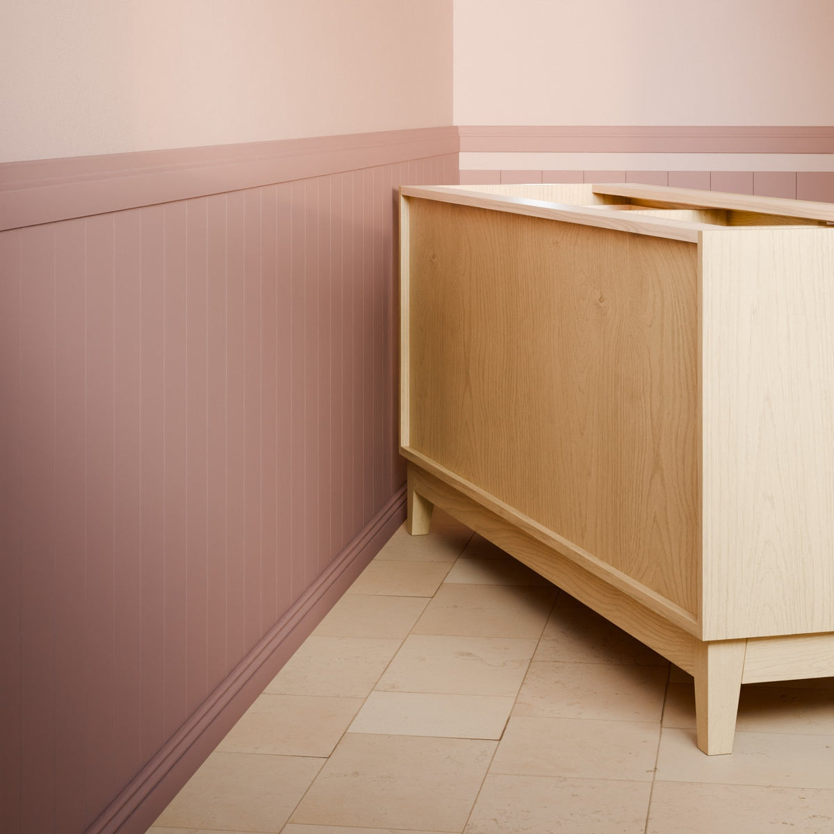A light natural wood cabinet or sideboard shown in a styled interior corner with pink-and-beige wainscoted walls and tiled floor.