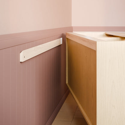 A close-up of a wall-mounted wooden trim piece on a dusty pink paneled wall next to a light wood cabinet or vanity unit.