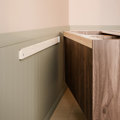 A close-up of a light-colored wooden wall-mounted fixture or trim piece installed on a paneled wall beside a dark wood cabinet or vanity in an interior room.