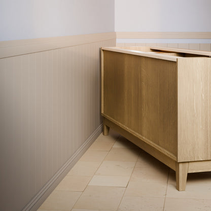 A light oak wood bathroom vanity cabinet shown in a real bathroom setting with beige tile flooring and paneled walls.