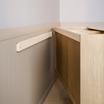 A light natural wood wall-mounted shelf or ledge installed on a beige paneled wall next to a wooden cabinet or counter in an indoor room setting.