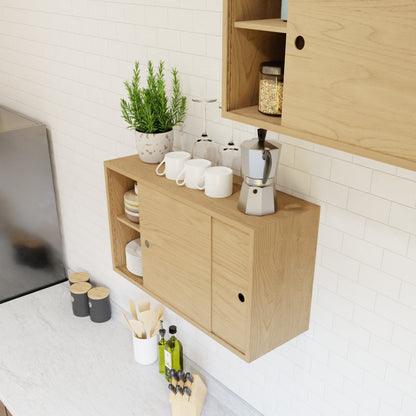 A compact kitchen counter features Krovel’s Hardwood Cupboard Shelf above, with light wood cabinets, a potted herb, cups, a stovetop espresso maker, glass jars, and utensils against a white tiled wall.
