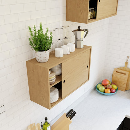 The Krovel Hardwood Cupboard Shelf is a wall-mounted cabinet with sliding doors for storing dishes and glasses. On top, it holds a potted plant, four wine glasses, four white mugs, and a moka pot above a bowl of fruit on the white countertop.