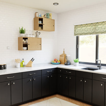Modern kitchen features black lower cabinets, a white countertop, Krovel's Hardwood Cupboard Shelf as upper storage, assorted utensils and appliances, a fruit bowl, and a window with a green patterned blind allowing natural light.