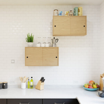 A minimalist kitchen features Krovel's Hardwood Cupboard Shelf displaying cups, a plant, a moka pot, and boxes. Below, the white tile backsplash and light wood cabinets frame a countertop with utensils, knives, olive oil, and fruit.
