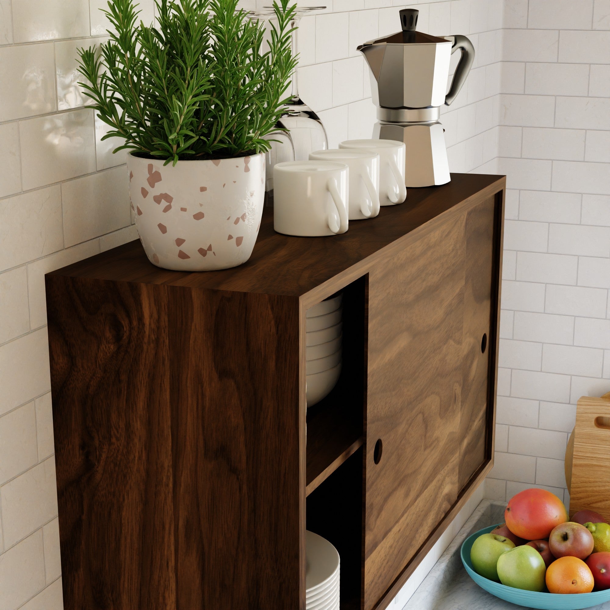The Krovel Hardwood Cupboard Shelf holds a potted plant, three white mugs, and a stovetop coffee maker. Bowls and plates are stored below. A bowl of fruit sits nearby on the counter, set against a white tile backsplash.