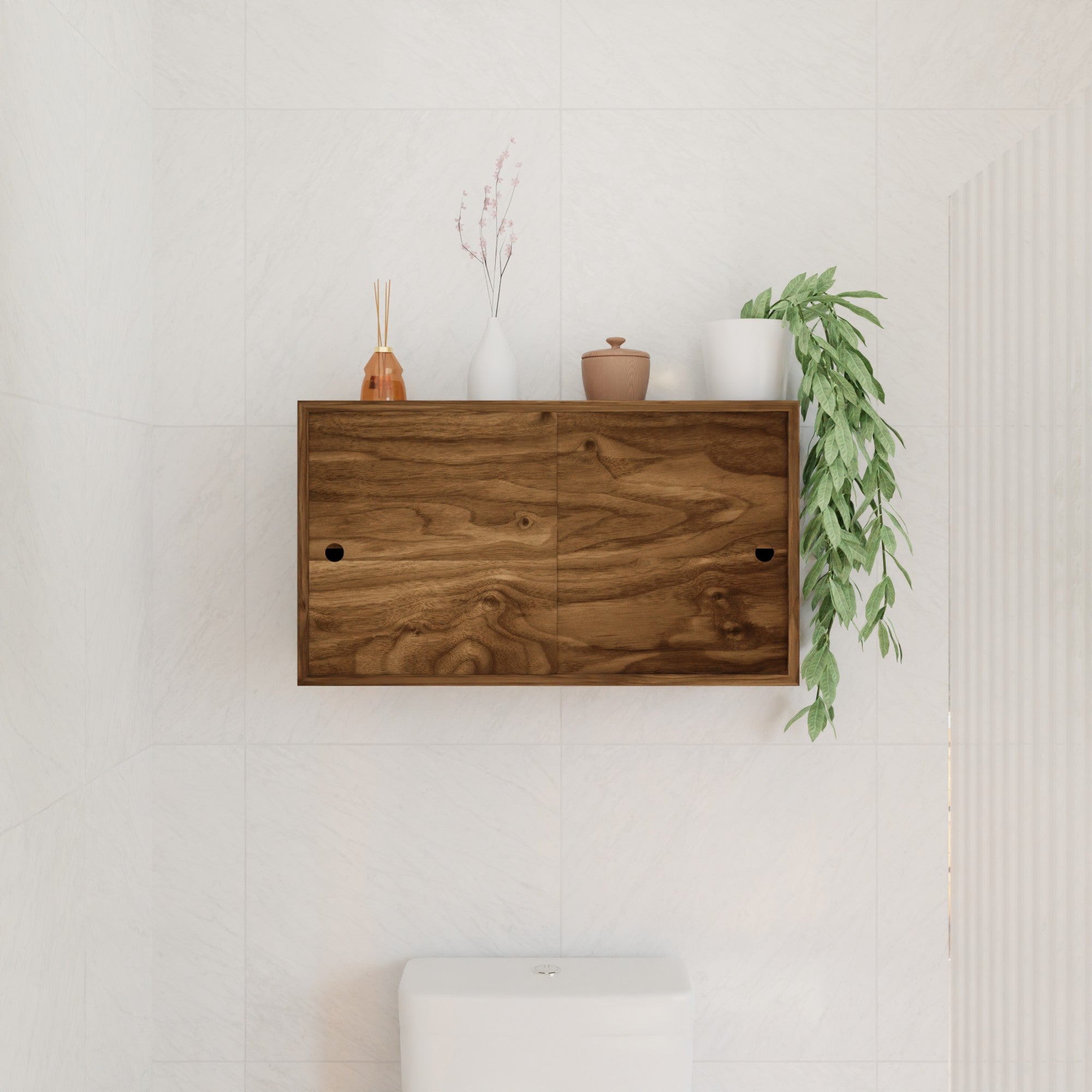 A Krovel Hardwood Cupboard Shelf is mounted above a white toilet, showcasing decor like a reed diffuser, white vase with branches, brown jar, white pot, and trailing green plant on a white-tiled wall.