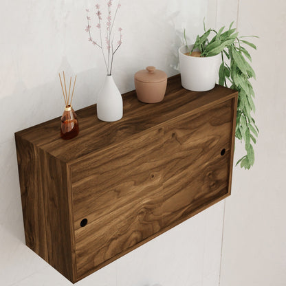 The Krovel Hardwood Cupboard Shelf is a wall-mounted cabinet with two doors, displayed against a light wall and styled with a pink floral vase, brown jar, white planter with greenery, and a reed diffuser on top.
