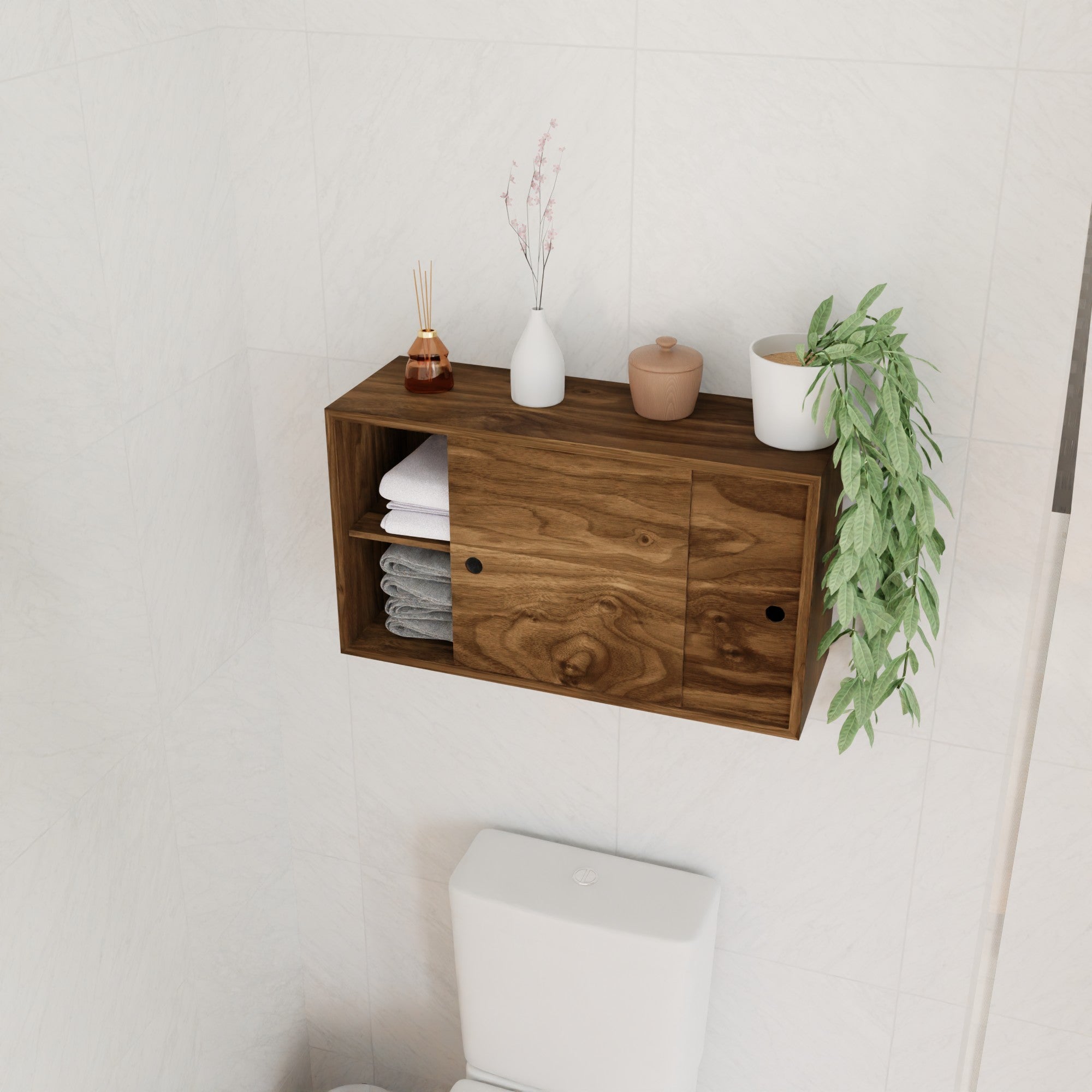 The Krovel Hardwood Cupboard Shelf is mounted above the toilet on a white-tiled wall, holding stacked towels, a reed diffuser, small vases, a lidded container, and a trailing green plant.