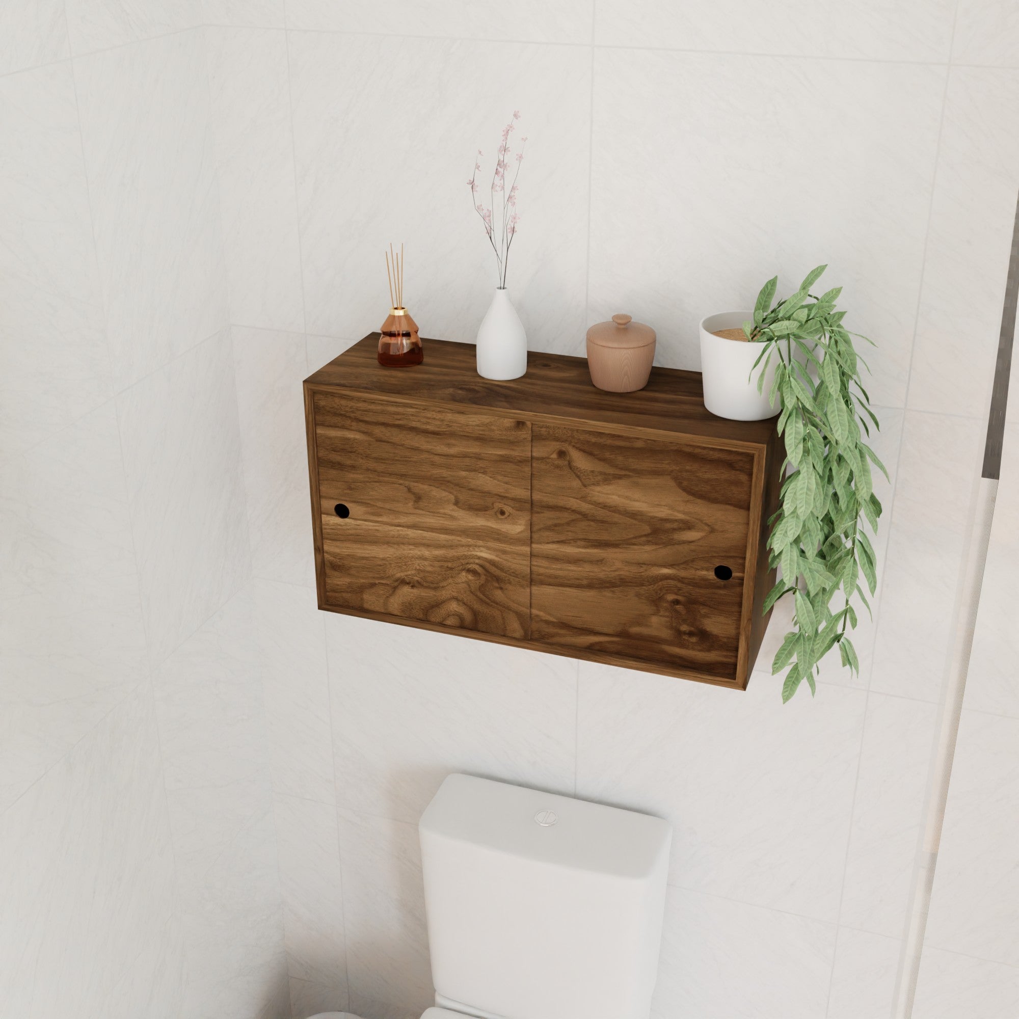 A Krovel Hardwood Cupboard Shelf with two doors is wall-mounted above a white toilet. Decorative items, such as vases, a diffuser, a ceramic container, and a trailing green plant are displayed on top of the cupboard.
