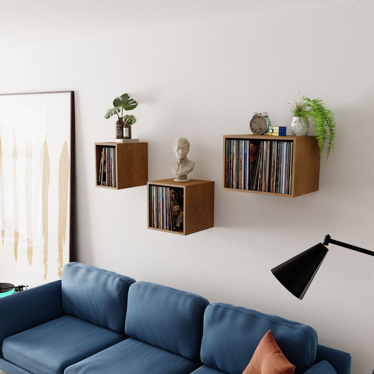 Three Krovel Record Storage Shelves in Cherry float above a blue sofa, displaying records, a small bust, plants, a clock, and decor. A black lamp and abstract art complete the modern living room's stylish look.