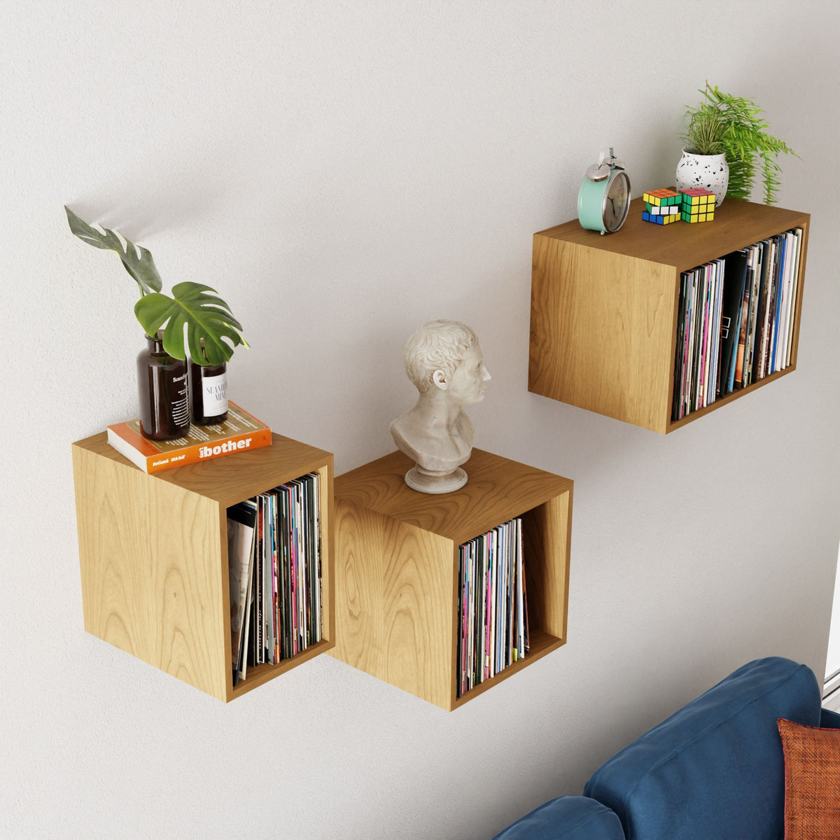 Three Krovel Record Storage Shelves in Cherry display vinyl records, books, décor, and more above a blue sofa against a light wall—offering stylish, versatile storage for your music and favorite items.