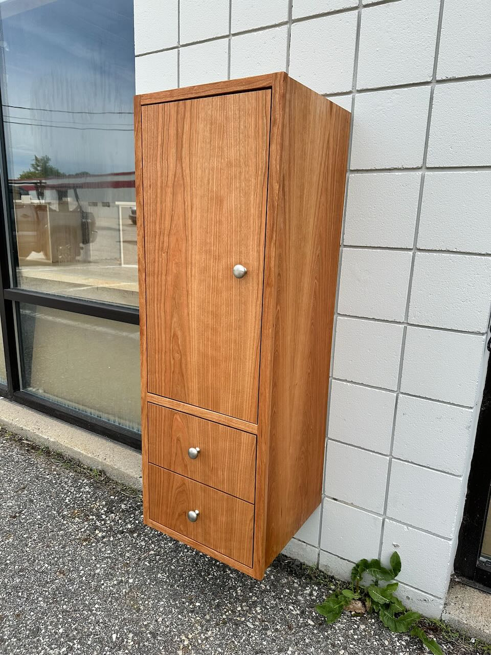 The Cherry Linen Cabinet - SAMPLE by Krovel Furniture Co. is a formaldehyde-free wooden cabinet featuring a door and two drawers, mounted on a white tiled wall outside near a window and placed on gravel next to a small green plant, making it an ideal choice for bathroom storage.