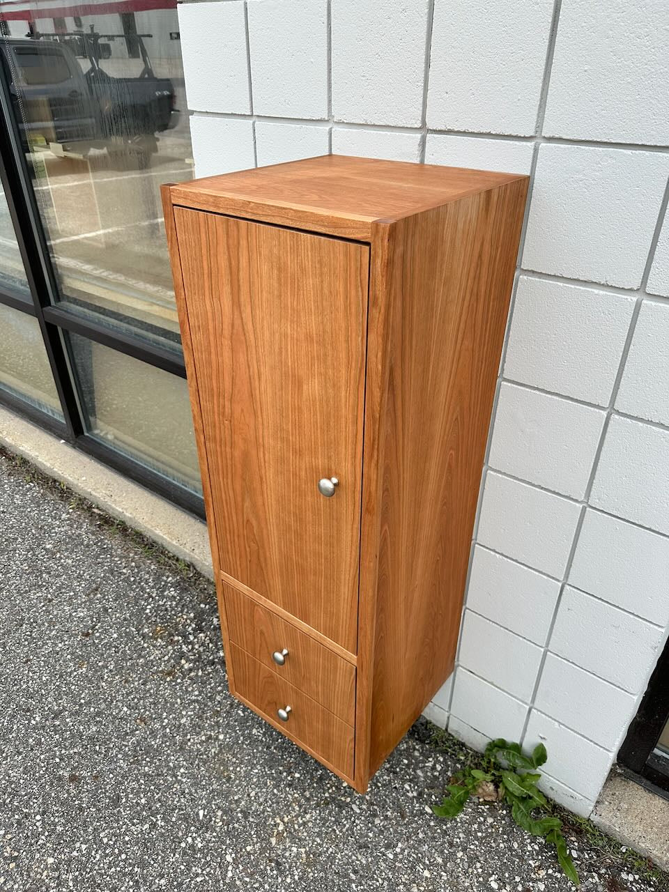 A formaldehyde-free Krovel Furniture Co. Cherry Linen Cabinet - SAMPLE with a door and two drawers stands against a white tiled wall outside on a gravel surface. A small green plant grows at the base of the bathroom storage cabinet.