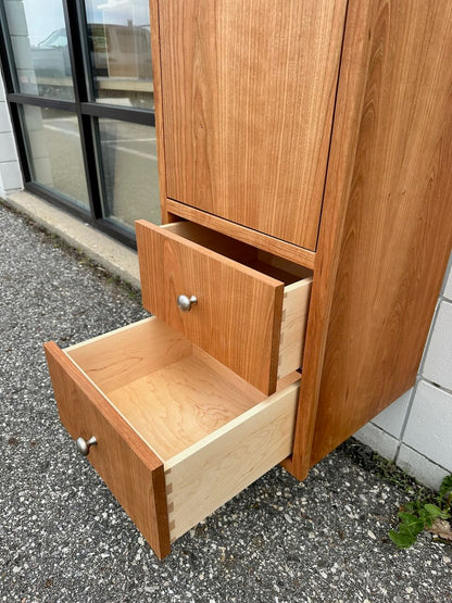 A formaldehyde-free Cherry Linen Cabinet - SAMPLE by Krovel Furniture Co., featuring two partially open drawers, sits on a gravel surface beside a white brick wall and large windows, making it an ideal bathroom storage cabinet.