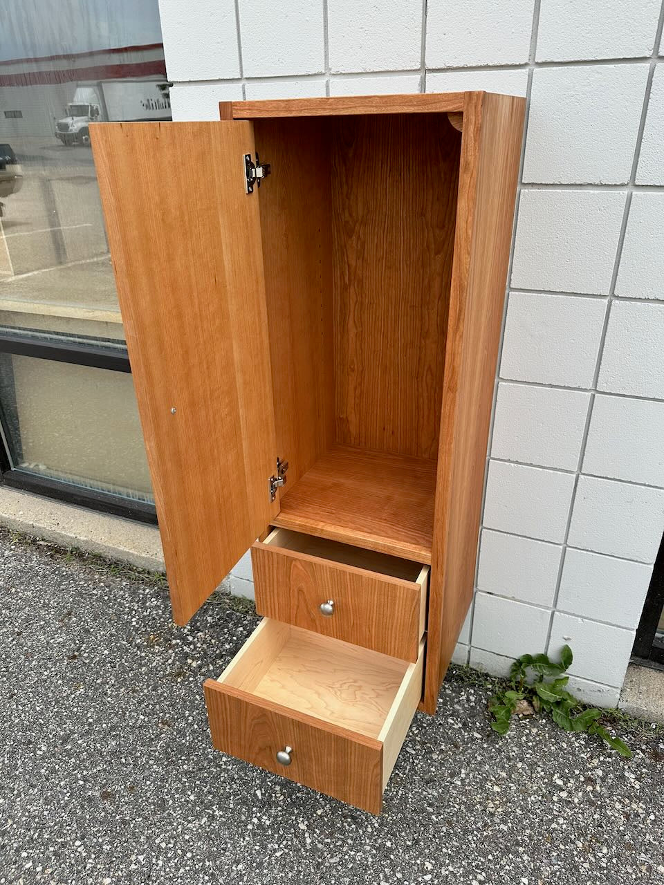 A formaldehyde-free Cherry Linen Cabinet - SAMPLE by Krovel Furniture Co. stands outside, its main door open and two lower drawers partially pulled out to reveal empty storage space—perfect as a bathroom storage cabinet or a stylish addition to your home.