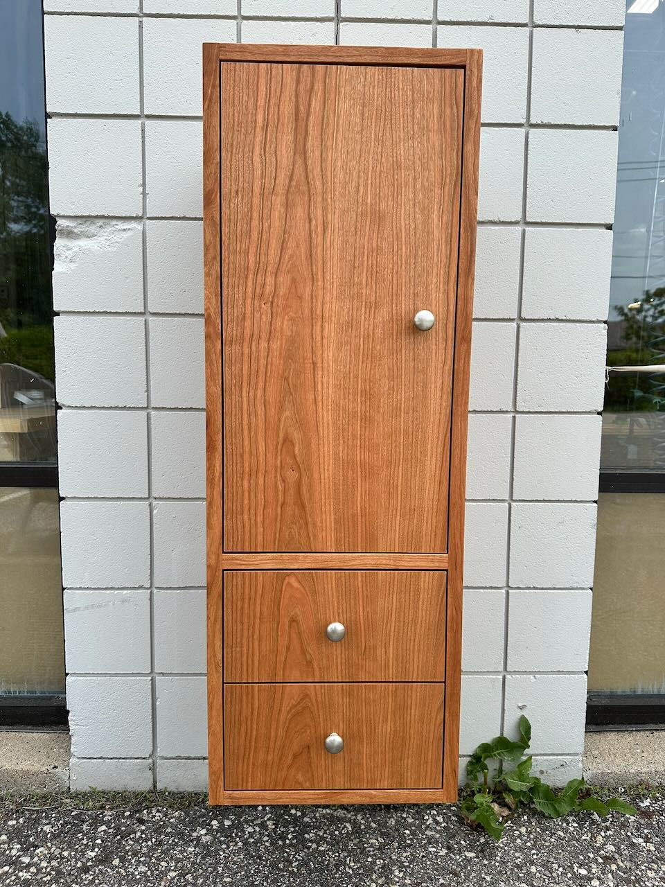 The Krovel Furniture Co. Cherry Linen Cabinet - SAMPLE is a tall, wooden bathroom storage cabinet featuring a single door on top and two drawers below, each adorned with a round silver knob. Shown placed outside against a white tiled wall with a small green plant at the bottom right, this formaldehyde-free cabinet combines functional storage with peace of mind for your home environment.