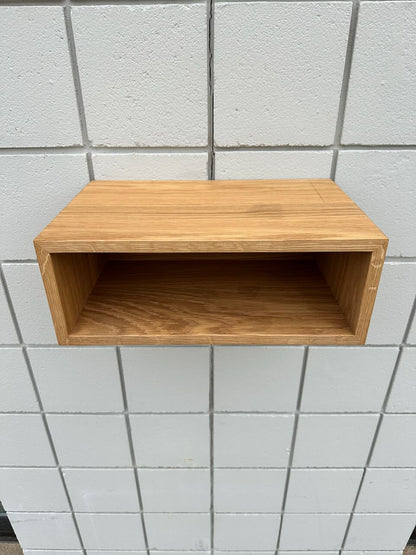 A simple, rectangular White Oak 16″ Nightstand Shelf - SECOND by Krovel Furniture Co. is mounted on a white tiled wall. Crafted from solid White Oak with a natural wood grain finish, this wall-mounted nightstand adds minimalist elegance to any space. The open-front shelf is empty, drawing attention to its clean and modern design.
