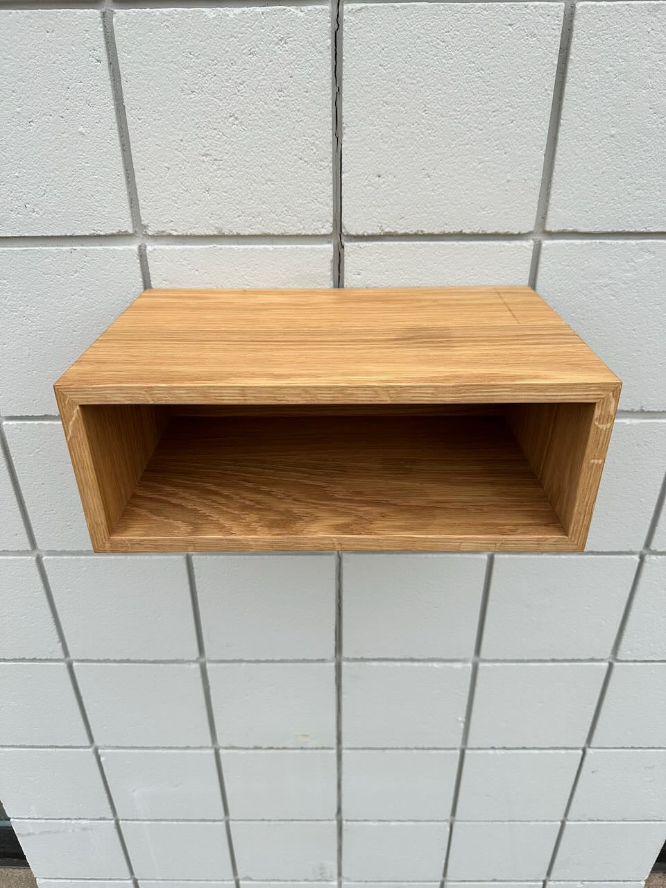 A simple, rectangular White Oak 16″ Nightstand Shelf - SECOND by Krovel Furniture Co. is mounted on a white tiled wall. Crafted from solid White Oak with a natural wood grain finish, this wall-mounted nightstand adds minimalist elegance to any space. The open-front shelf is empty, drawing attention to its clean and modern design.