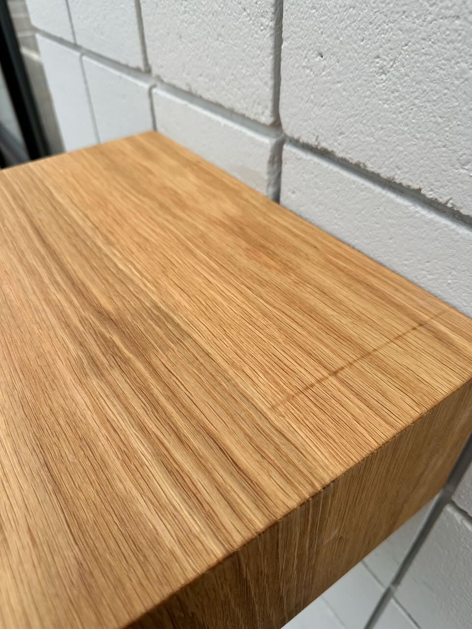 A close-up of the corner of the White Oak 16″ Nightstand Shelf – SECOND by Krovel Furniture Co. showcases the natural grain of its solid White Oak construction, set against a backdrop of white tiled wall. The wood’s surface appears smooth and expertly finished.