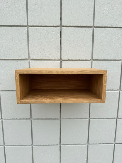 A White Oak 16″ Nightstand Shelf - SECOND by Krovel Furniture Co. is mounted on a wall of white square tiles. The rectangular shelf, crafted from solid White Oak, has its open side facing forward and is empty. The image is centered on both the floating nightstand shelf and the grout lines of the tiled wall.