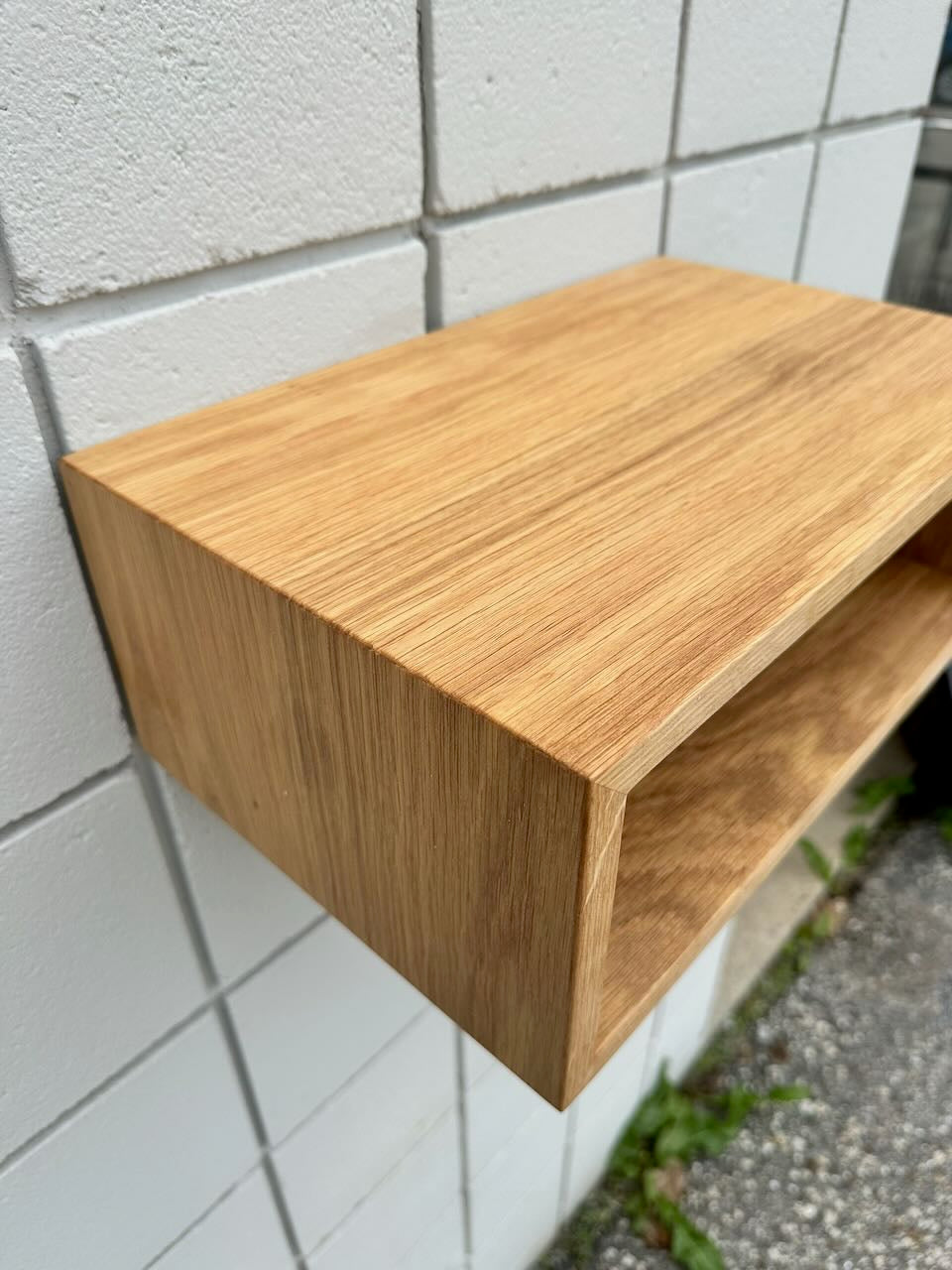 A Krovel Furniture Co. White Oak 16″ Nightstand Shelf - SECOND, featuring a rectangular design, is mounted on a tiled white wall above an outdoor area with visible grass and pavement.