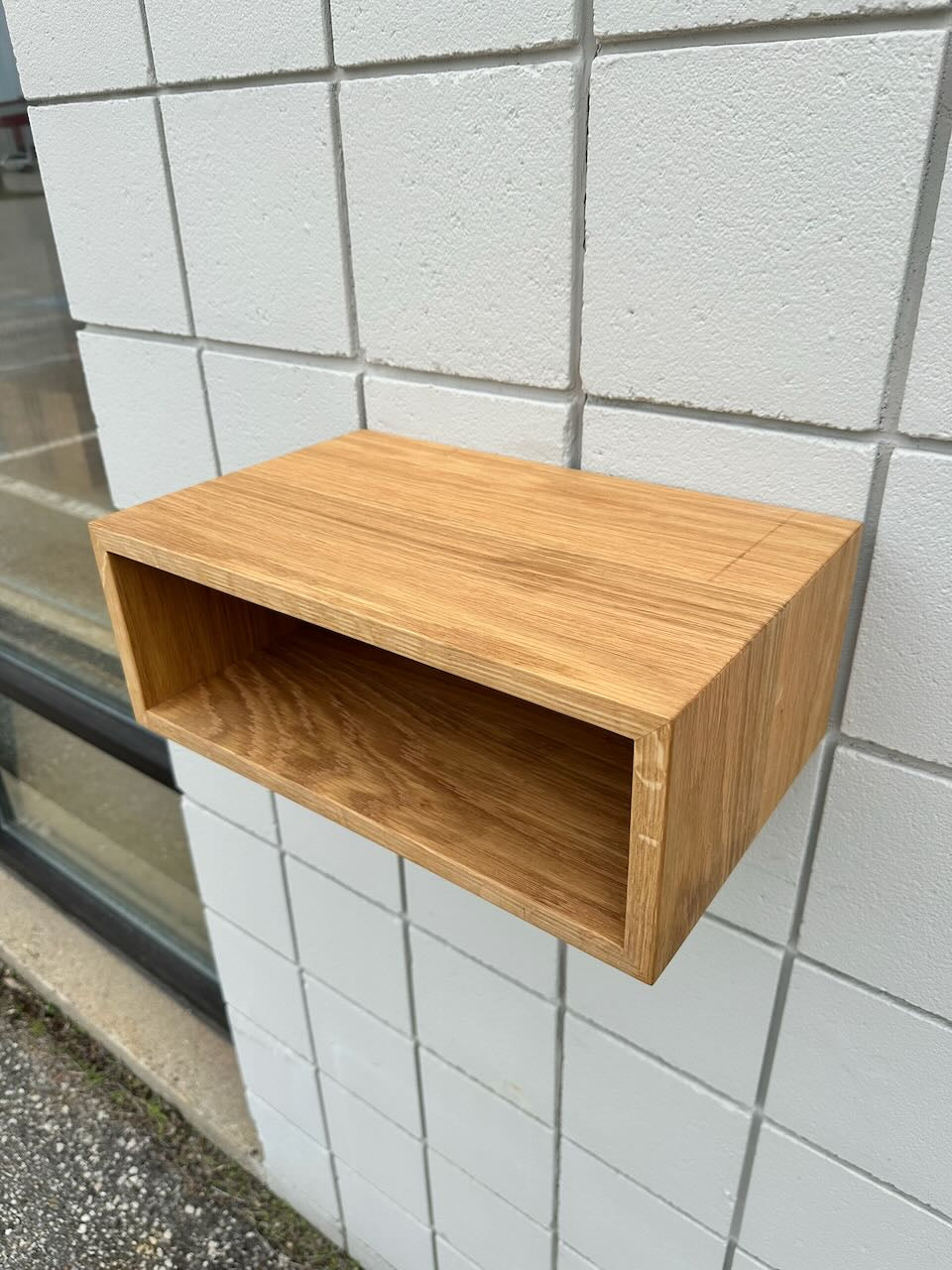 A compact, rectangular White Oak 16″ Nightstand Shelf - SECOND by Krovel Furniture Co. is mounted on a white tiled wall. Crafted from solid White Oak, the shelf features an open front and a simple, minimalist design that evokes the look of a floating nightstand. In the background, part of a window reveals an outdoor view.