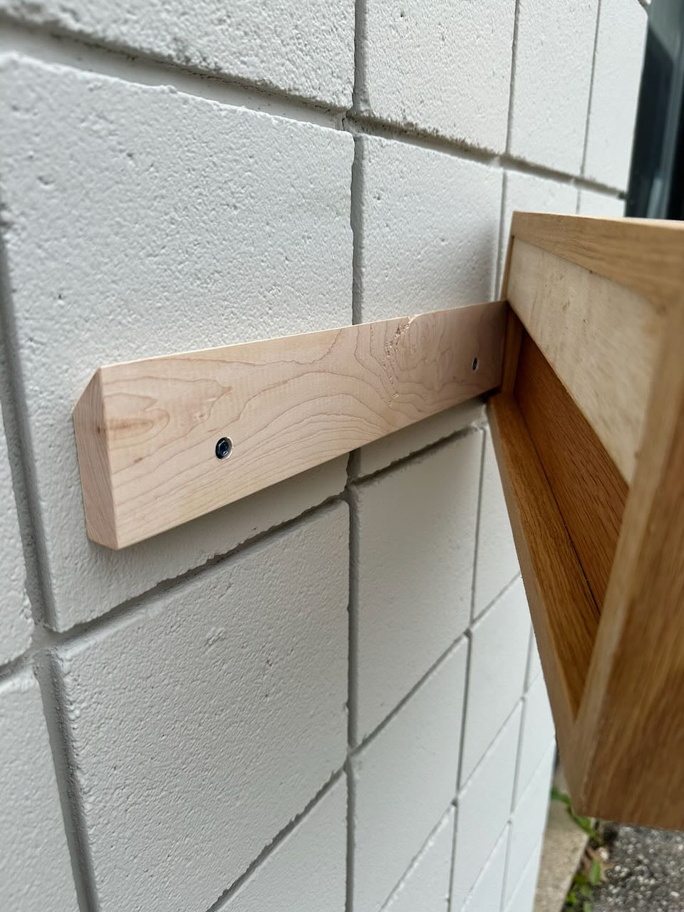 A White Oak 16″ Nightstand Shelf - SECOND by Krovel Furniture Co. is securely mounted to a white tiled wall using a solid White Oak French cleat, with the edge of the wooden shelf resting on it—clearly demonstrating the sturdy wall-mounting method often used for floating nightstands.