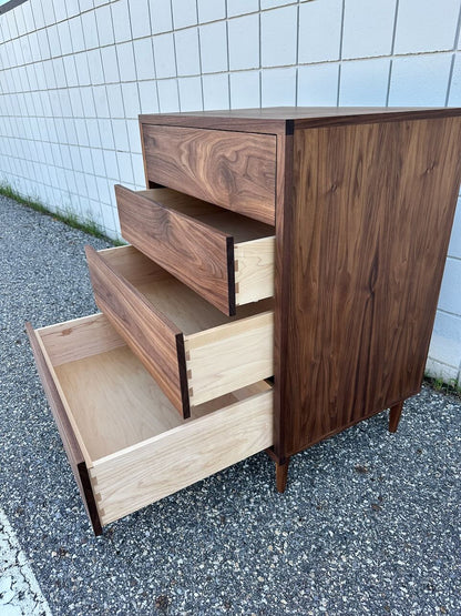 A 4 Drawer Dresser in Walnut by Krovel Furniture Co. stands on an outdoor pavement beside a white tiled wall, with three of its drawers open to reveal light-colored interiors. Expertly crafted from solid hardwood and finished with a zero-VOC oil finish, this dresser combines natural elegance with eco-friendly design.