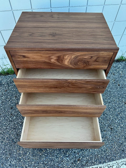A solid walnut four-drawer dresser by Krovel Furniture Co., with drawers open to reveal empty, light-colored interiors, is placed on pebbled ground against a white tiled wall. Finished with a zero-VOC oil for a natural and eco-friendly look.