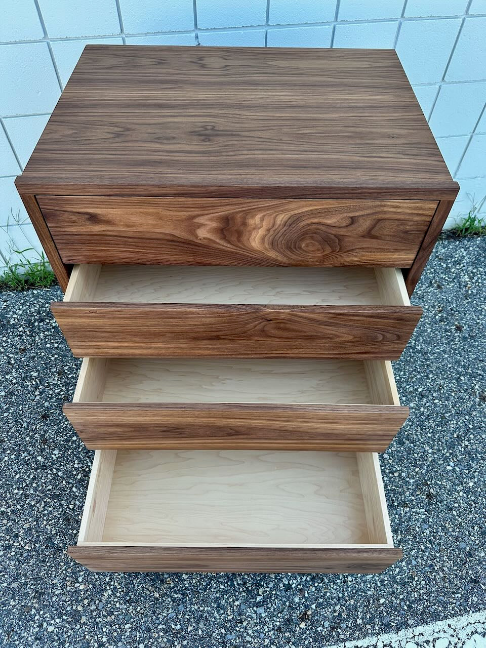 A solid walnut four-drawer dresser by Krovel Furniture Co., with drawers open to reveal empty, light-colored interiors, is placed on pebbled ground against a white tiled wall. Finished with a zero-VOC oil for a natural and eco-friendly look.
