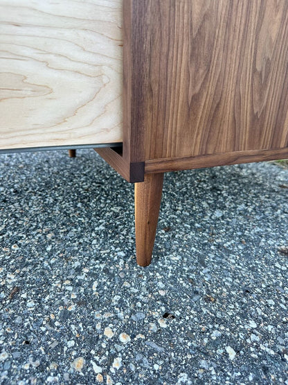 Close-up of a solid hardwood leg and corner of the 4 Drawer Dresser in Walnut - SAMPLE by Krovel Furniture Co., highlighting two-tone wood grain and precise joinery with a zero-VOC oil finish, pictured on textured asphalt.