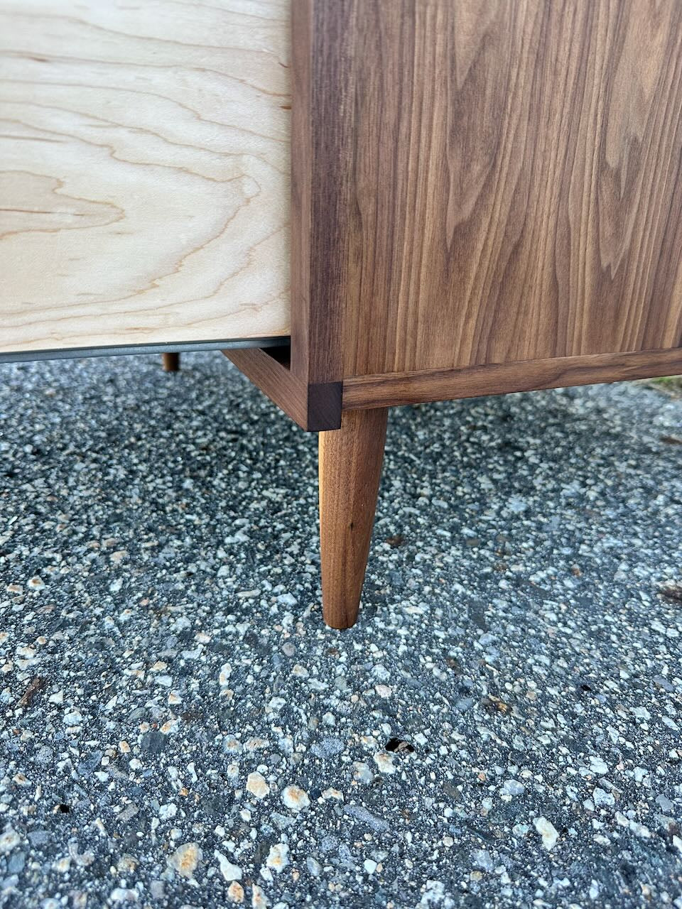Close-up of a solid hardwood leg and corner of the 4 Drawer Dresser in Walnut - SAMPLE by Krovel Furniture Co., highlighting two-tone wood grain and precise joinery with a zero-VOC oil finish, pictured on textured asphalt.