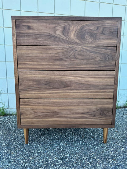 The 4 Drawer Dresser in Walnut - SAMPLE by Krovel Furniture Co. is crafted from solid hardwood walnut with four drawers, showcasing a rich wood grain pattern and finished in a zero-VOC oil. It stands on short legs outdoors against a tiled wall and gravel ground.
