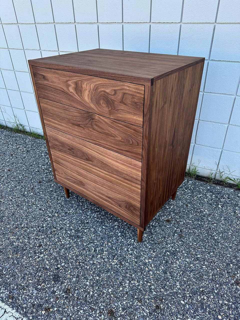 A mid-century modern 4 Drawer Dresser in Walnut by Krovel Furniture Co. stands on a gravel surface in front of a white tiled wall. Expertly crafted from solid hardwood, this piece features a rich natural grain pattern, tapered legs, and an eco-friendly zero-VOC oil finish.