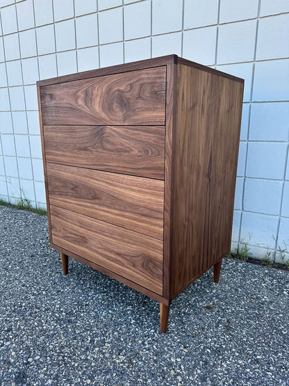 The 4 Drawer Dresser in Walnut by Krovel Furniture Co., a mid-century modern piece crafted from solid hardwood with four drawers, tapered legs, and a zero-VOC oil finish, sits on a gravel surface against a white tiled wall.