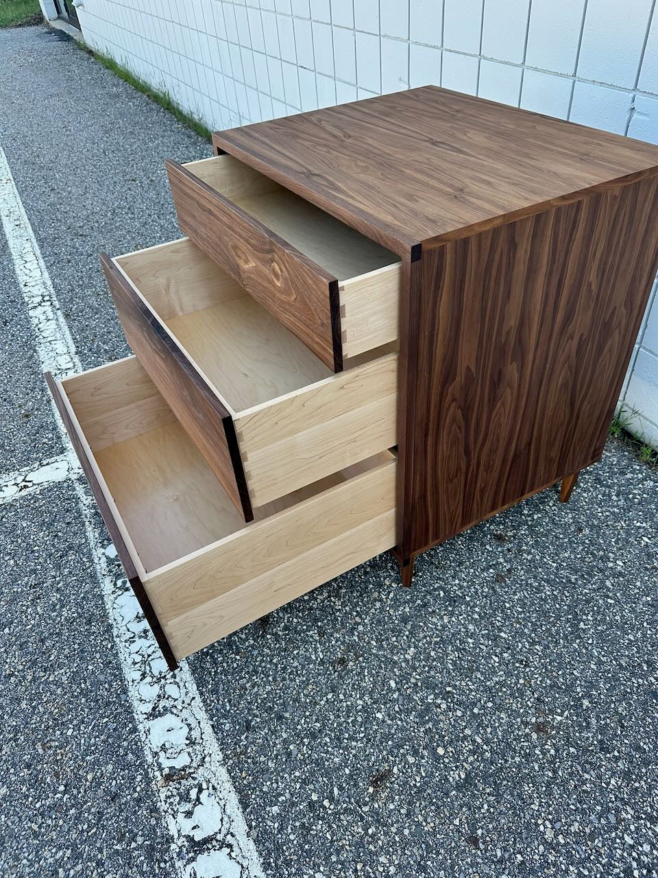 The 3 Drawer Dresser in Walnut - SECOND by Krovel Furniture Co. stands on a gravel surface beside a white-tiled building, its three drawers partially open to reveal a light wood interior contrasting with the darker walnut exterior finished in zero-VOC.